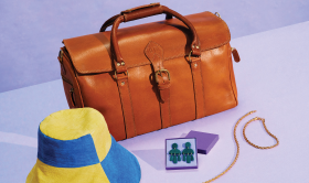 Fotografia przedstawiająca brązową, skórzaną torbę podróżną, kapelusz z żółtymi i niebieskimi panelami, zestaw ciemnoszmaragdowych kolczyków w fioletowym pudełku na biżuterię oraz złoty warkoczowy naszyjnik i zestaw bransoletek. Przedmioty znajdują się na lawendowym tle z małym logo Etsy czarną czcionką w lewym górnym rogu.