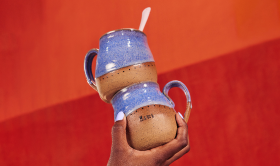 Photograph of a hand holding two stacked handmade ceramic mugs with a natural brown clay base dipped in a light blueish purple glaze on an orange red background with a small Etsy logo in white font in the top left corner