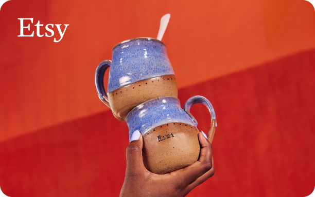 Foto einer Hand, die zwei gestapelte handgefertigte Keramikbecher mit einer Basis aus natürlichem braunem Ton hält, die in eine hellblau-violette Glasur getaucht wurden, auf einem orange-roten Hintergrund mit einem kleinen Etsy-Logo in weißer Schrift in der oberen linken Ecke
