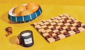 Still life photograph of a chequerboard with wooden pieces on a yellow tabletop. To the left, there’s a candle with the label “L’APOTHECARY” and a cork base. In the background, a blue striped bowl filled with citrus fruit.