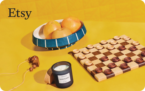 黄色のテーブルの上にある木のピースが置かれたチェッカー盤の写真左には、「L’APOTHECARY」のラベルが貼られたキャンドルとコルクベースがあります。背景には、柑橘類でいっぱいの青いストライプが入ったボウルがあります。