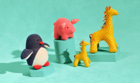 Still life photograph of plush toy animals on a teal background. On the left, a penguin with a white belly, black body and red feet. In the centre, a pink pig with darker pink details sits atop a teal block. To the right, two giraffes – one large with yellow fabric and orange spots, and a smaller one with lighter yellow and orange spots – stand near each other, with the smaller giraffe in front of a shorter teal block.